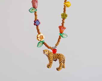 May include: A beaded necklace featuring a cheetah pendant. The necklace has a brown beaded chain with colorful flower and leaf accents. The cheetah pendant is tan with black spots and is connected to the chain with a red heart charm.