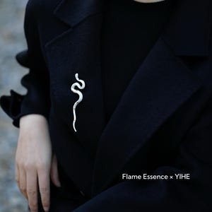 May include: A silver-colored snake brooch pinned to a black coat. The brooch is a stylized representation of a snake, with a curved, sinuous design. The text "Flame Essence x YIHE" is visible.