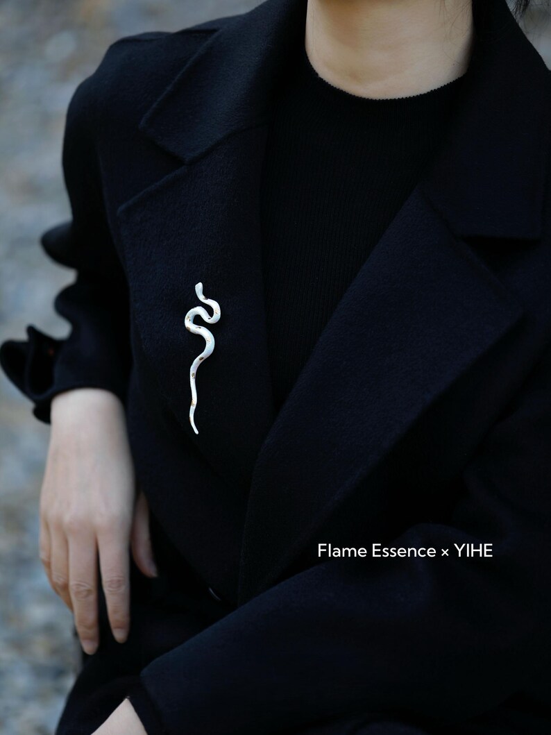 May include: A silver-colored snake brooch pinned to a black coat. The brooch is a stylized representation of a snake, with a curved, sinuous design. The text "Flame Essence x YIHE" is visible.