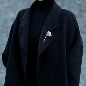 K&ouml;nnte beinhalten: Eine silberfarbene Brosche in Form eines Mantarochens ist an einem schwarzen Mantel befestigt. Die Brosche hat ein detailliertes Design mit strukturiertem K&ouml;rper und einem langen, gebogenen Schwanz. Der Mantel ist dunkel und die Brosche bildet einen Kontrast.