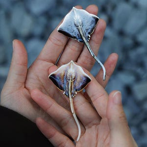 K&ouml;nnte beinhalten: Zwei Miniatur-Stechrochen-Skulpturen. Die Rochen haben eine wei&szlig;e Unterseite, eine dunkelblaue und schwarze Oberseite und einen langen, gebogenen Schwanz. Die Skulpturen werden in offenen H&auml;nden vor einem verschwommenen Hintergrund gehalten.