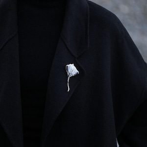 K&ouml;nnte beinhalten: Eine silberfarbene Brosche in Form eines stilisierten Rochens ist an einem schwarzen Mantel befestigt. Die Brosche hat ein strukturiertes, fl&uuml;gelartiges Design und einen schlanken Stiel. Der Mantel ist Teil eines schwarzen Outfits, mit einem schwarzen Rollkragen unter dem Revers.