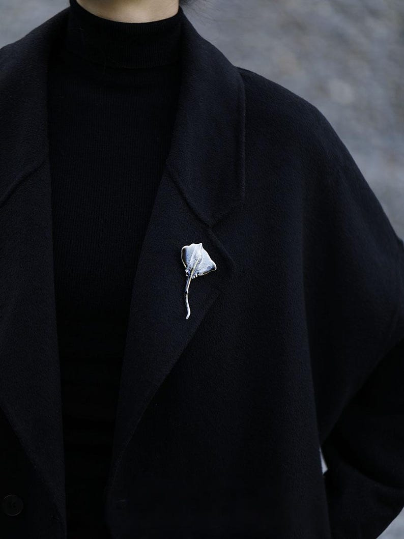 K&ouml;nnte beinhalten: Eine silberfarbene Brosche in Form eines stilisierten Rochens ist an einem schwarzen Mantel befestigt. Die Brosche hat ein strukturiertes, fl&uuml;gelartiges Design und einen schlanken Stiel. Der Mantel ist Teil eines schwarzen Outfits, mit einem schwarzen Rollkragen unter dem Revers.