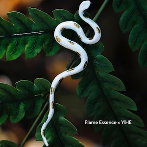 May include: A white snake figurine with gold-colored markings, resting on a bed of green fern leaves. The snake is in a curved pose, with its head raised. The text "Flame Essence x YIHE" is visible in the lower right corner.