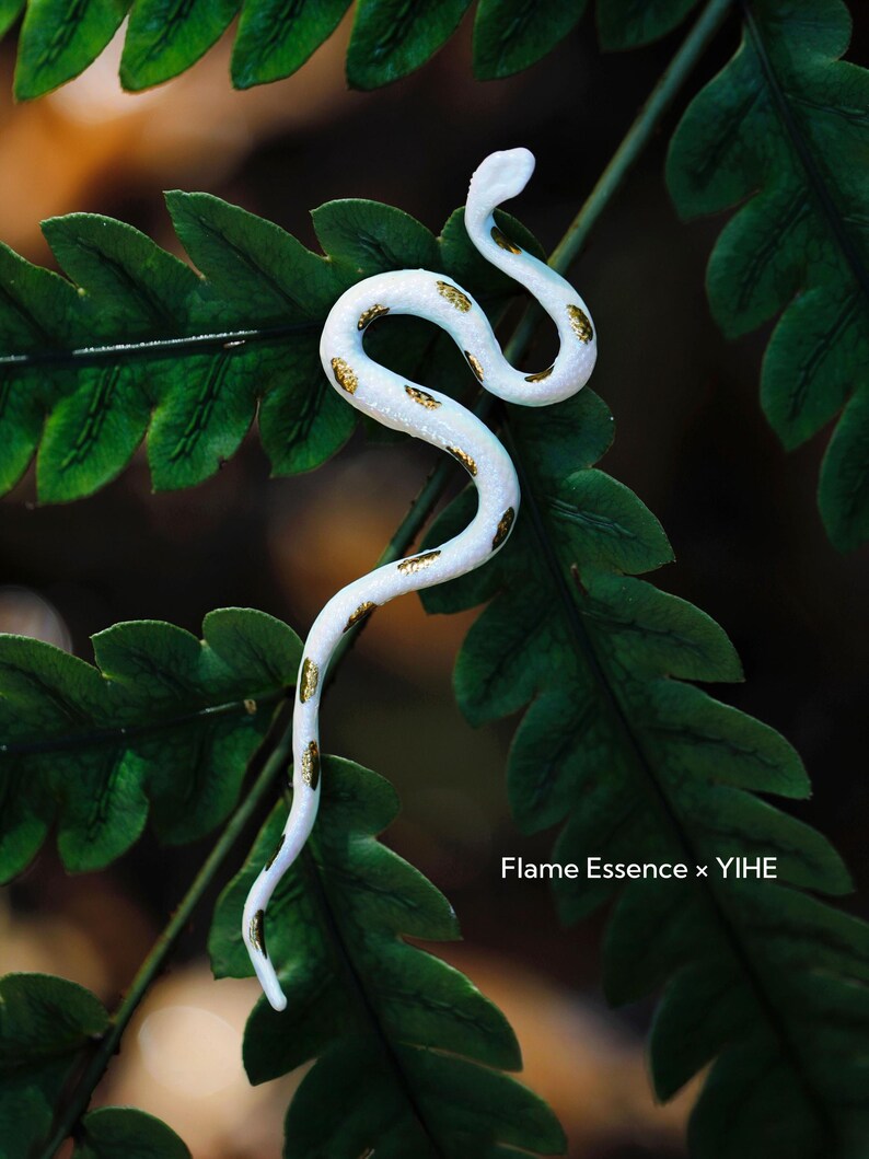 May include: A white snake figurine with gold-colored markings, resting on a bed of green fern leaves. The snake is in a curved pose, with its head raised. The text "Flame Essence x YIHE" is visible in the lower right corner.