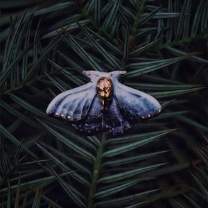 Puede incluir: Primer plano detallado de un broche con forma de polilla, con cabeza y detalles dorados. Las alas de la polilla presentan un degradado de azul y blanco con puntos dorados. El broche se encuentra sobre un fondo de follaje verde oscuro. El texto "Flame Essence x YIHE" está en la parte inferior.