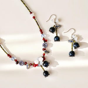 May include: A delicate necklace and matching earrings set. The necklace and earrings feature a variety of beads in red, white, blue, and purple. The earrings have a bell-shaped charm. The jewelry is on a white background.