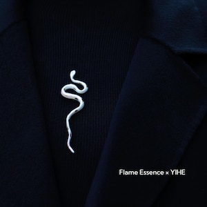 May include: A silver-colored snake-shaped brooch pinned to a black sweater and coat. The brooch has a textured, organic design. The text "Flame Essence x YIHE" is visible in the lower right corner.