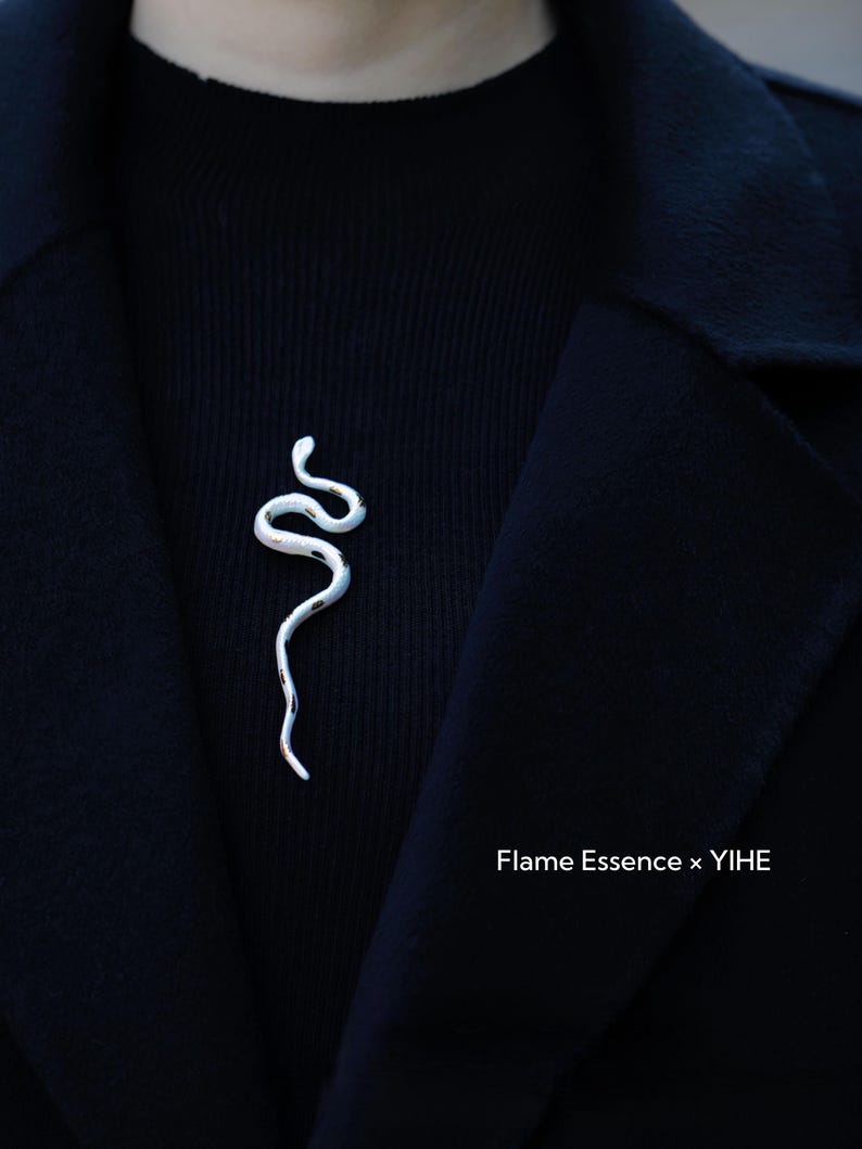 May include: A silver-colored snake-shaped brooch pinned to a black sweater and coat. The brooch has a textured, organic design. The text "Flame Essence x YIHE" is visible in the lower right corner.