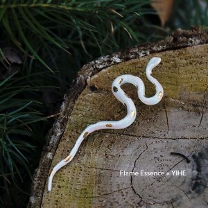May include: A white, serpentine figurine with gold leaf accents, resting on a weathered wooden surface. The snake-like sculpture is detailed and elegant, with a subtle iridescent sheen. The background features green foliage and the text "Flame Essence x YIHE".
