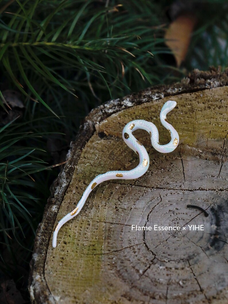May include: A white, serpentine figurine with gold leaf accents, resting on a weathered wooden surface. The snake-like sculpture is detailed and elegant, with a subtle iridescent sheen. The background features green foliage and the text "Flame Essence x YIHE".