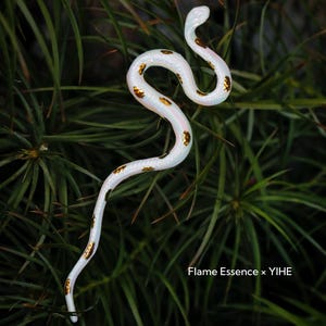 May include: A white snake figurine with gold-colored spots, posed against a backdrop of green foliage. The snake is slender and curved, with a slight iridescent sheen. The text "Flame Essence x YIHE" is visible.