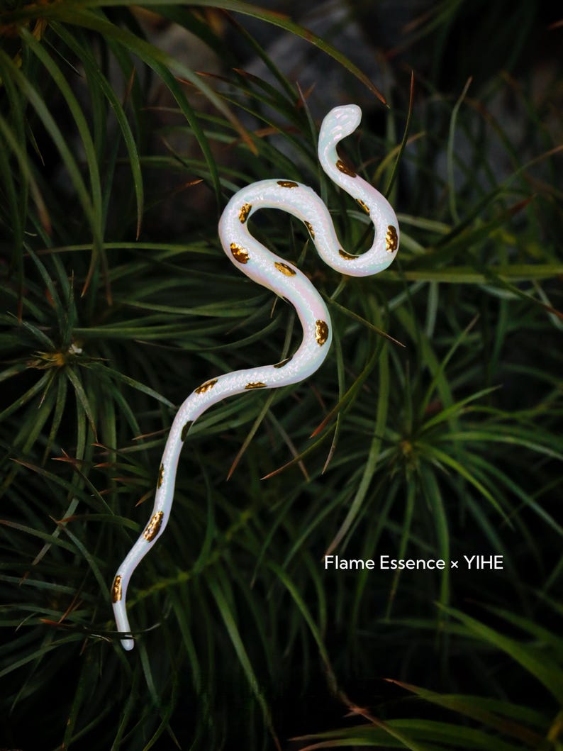 May include: A white snake figurine with gold-colored spots, posed against a backdrop of green foliage. The snake is slender and curved, with a slight iridescent sheen. The text "Flame Essence x YIHE" is visible.