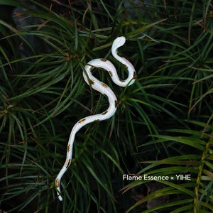 May include: A white snake figurine with gold-colored markings, coiled among green foliage. The snake is the focal point, with the text "Flame Essence x YIHE" visible in the lower right corner.