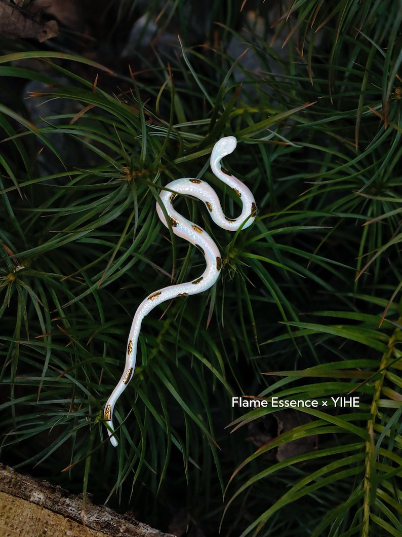 May include: A white snake figurine with gold-colored markings, coiled among green foliage. The snake is the focal point, with the text "Flame Essence x YIHE" visible in the lower right corner.