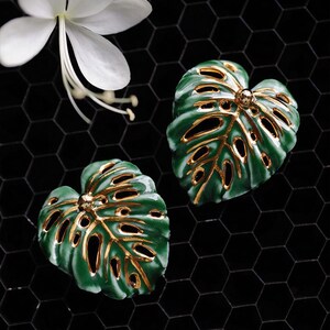 May include: A pair of green and gold leaf-shaped earrings with a glossy finish. The earrings feature a detailed design with cut-out sections, set against a black honeycomb background. A white flower is in the upper left corner. The text "Flame Essence x YIHE" is at the bottom.