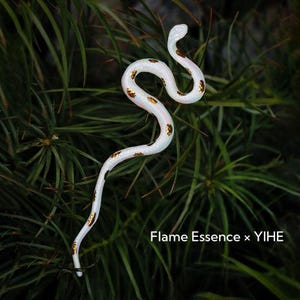 Könnte beinhalten: Ein weißes, schlangenartiges Deko-Objekt mit goldenen Punkten, eingebettet in grünes Blattwerk. Der Text "Flame Essence x YIHE" ist unten im Bild zu sehen.