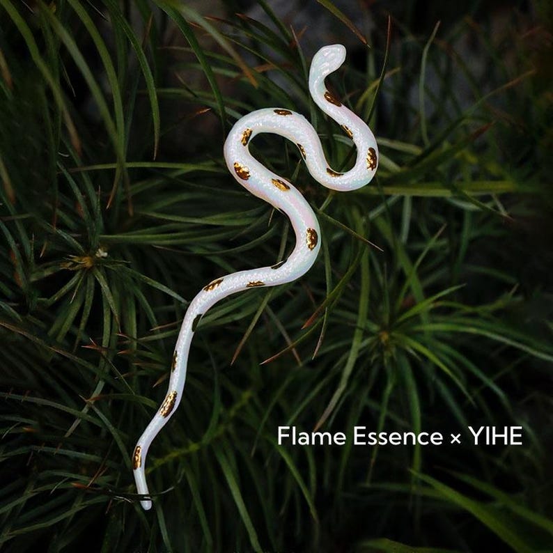 May include: A white, serpentine decorative object with gold-colored spots, resting on a bed of green foliage. The text "Flame Essence x YIHE" is visible at the bottom of the image.