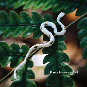 May include: A white and gold-spotted snake figurine rests on a vibrant green fern. The snake is curled, with its head raised, showcasing the intricate details of its design. The image is a close-up, highlighting the textures and colors of the snake and foliage.