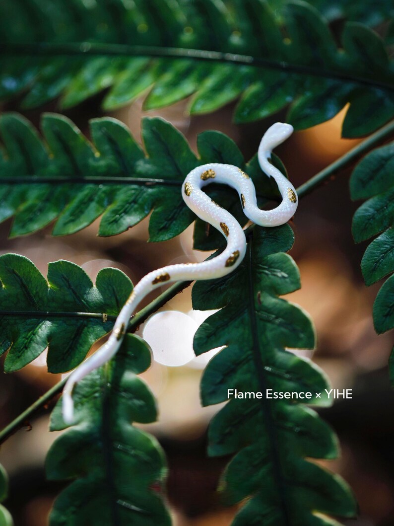 May include: A white and gold-spotted snake figurine rests on a vibrant green fern. The snake is curled, with its head raised, showcasing the intricate details of its design. The image is a close-up, highlighting the textures and colors of the snake and foliage.