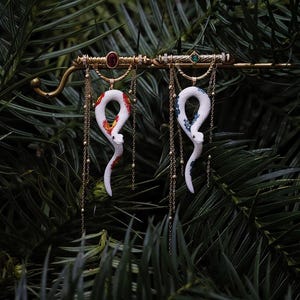 Op de afbeelding: Gouden sieraad met twee witte slangenhangers met kleurrijke accenten. De hangers hangen aan een decoratieve staaf versierd met edelstenen, tegen een achtergrond van groen gebladerte.