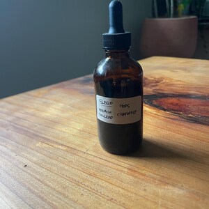 Pode incluir: Uma garrafa de vidro castanho com um conta-gotas preto cheia de um líquido escuro. Um rótulo branco na garrafa diz "SLEEP" com os ingredientes "VERVAIN, SKULLCAP, HOPS, CHAMOMILE". A garrafa está sobre uma superfície de madeira.