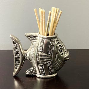 May include: A silver-toned metal toothpick holder shaped like a fish with textured scales and fins. The hollow body of the fish serves as a container for a cluster of light-colored wooden toothpicks that protrude from the top opening.