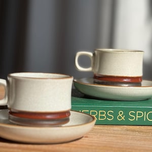 Puede incluir: Dos tazas de café color crema con bandas marrones y platillos a juego. Las tazas tienen un diseño simple y clásico. Las tazas y platillos están sobre una superficie de madera. El texto "HERBS & SPICES" es visible en un libro verde.