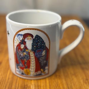 May include: White ceramic mug featuring a festive illustration of Santa Claus in a red coat, holding a Christmas tree and gifts. The mug has a curved handle and a gold-toned border around the Santa image. The mug is a perfect holiday gift.