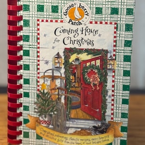 May include: A spiral-bound cookbook titled "Coming Home for Christmas" by Gooseberry Patch. The cover features a red door with a Christmas wreath, a small Christmas tree, and a green and white plaid pattern. The book includes recipes and holiday ideas.