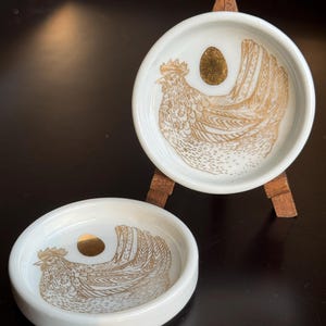 May include: Two white ceramic circular trinket dishes featuring a gold-toned illustration of a hen and a single gold egg. One dish rests flat while the other is displayed upright on a small wooden easel stand, showcasing the detailed line art.