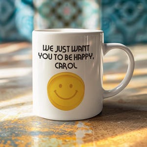 May include: White ceramic mug with a curved handle. The mug features the text "WE JUST WANT YOU TO BE HAPPY, CAROL" in black, above a yellow smiley face design.