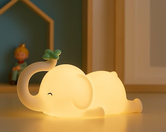 May include: A soft, glowing elephant-shaped night light. The light is a warm yellow, with a curved trunk and a small green leaf detail. The elephant is lying down, with a gentle smile.