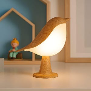 Op de afbeelding: Een houten vogelvormige tafellamp met een warm, wit gloeiend licht. De lamp heeft een lichtbruine houten behuizing en een witte, afgeronde buik. Hij staat op een houten voet, wat een charmant en grillig ontwerp creëert. Een decoratieve lamp.