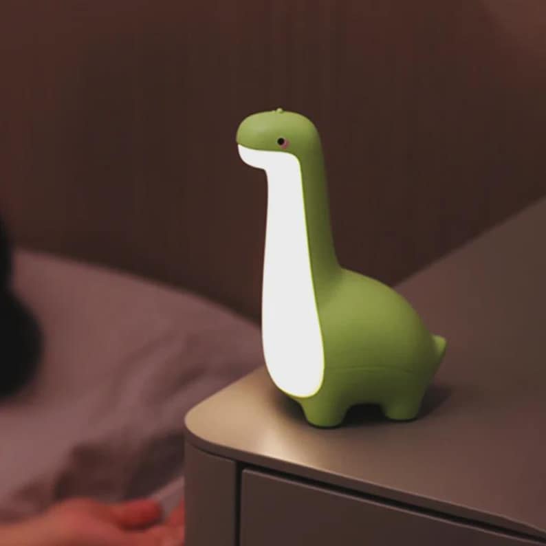 May include: A green dinosaur-shaped night light with a long neck and a glowing white center. The lamp is made of a smooth material and has a small pink detail on its head. It is placed on a gray nightstand.
