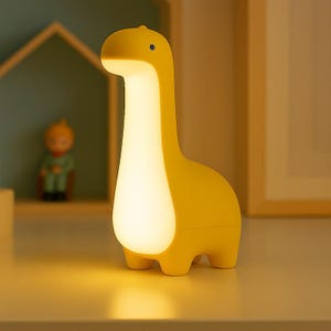 May include: A yellow dinosaur-shaped night light with a glowing white center. The lamp has a long neck, a small head with a blue dot, and four stubby legs. The lamp is on a white surface.