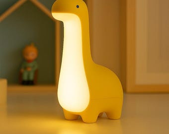 May include: A yellow dinosaur-shaped night light with a glowing white center. The lamp has a long neck, a small head with a blue dot, and four stubby legs. The lamp is on a white surface.