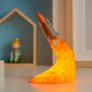 Może przedstawiać: Dekoracyjna lampa w kształcie startującego wahadłowca kosmicznego. Wahadłowiec jest pomarańczowy i szary, z jasnopomarańczowym efektem płomienia u podstawy. Lampa ma wyglądać jak startująca rakieta.