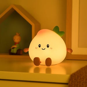 以下が含まれることがあります： 笑顔の顔、小さな腕と脚が付いた、光る桃の形をしたナイトライト。ライトはピンク色のトップ、緑の葉、茶色の腕と脚をしています。ナイトライトは黄色の表面に置かれています。