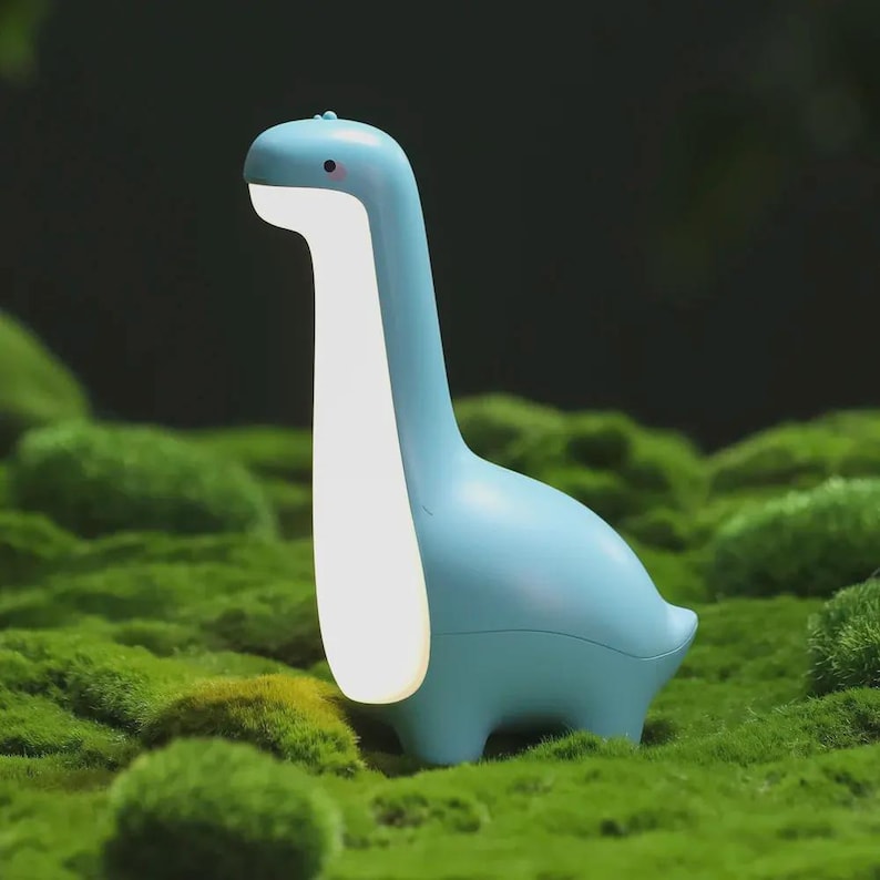 May include: A light blue dinosaur-shaped lamp with a long neck and a glowing white light. The lamp has a small red dot for an eye and is set against a backdrop of green moss. A whimsical and decorative lighting fixture.