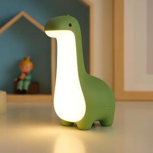 May include: A green dinosaur-shaped night light with a glowing white center. The lamp has a long neck and a small head, with a light shining from the top. The lamp is on a white surface.