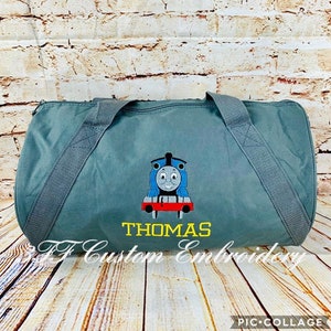 May include: A gray canvas duffel bag with a blue and red Thomas the Tank Engine embroidered design and the name "THOMAS" in yellow lettering.