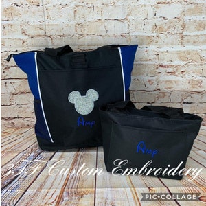 May include: Two black tote bags with blue accents. The larger bag has a silver glitter Mickey Mouse head and the name "Amy" embroidered in blue. The smaller bag has the name "Amy" embroidered in blue.