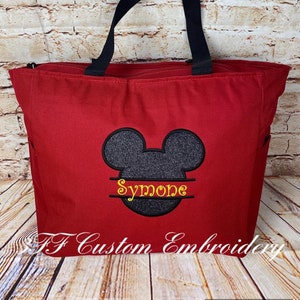 Puede incluir: Bolsa de mano roja con asas negras y una cabeza de Mickey Mouse negra y gris con el nombre "Simone" bordado en amarillo en la parte inferior.