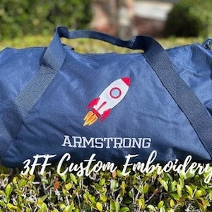 May include: A navy blue duffel bag with a white embroidered rocket ship and the name "ARMSTRONG" underneath. The bag also has the text "3FF Custom Embroidery" on the bottom.