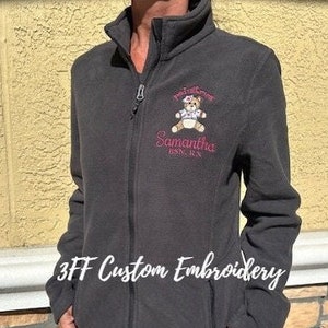 May include: A black fleece jacket with a zipper closure and a pink embroidered design featuring a teddy bear with a pink bow and the text "Pawfectly Cute Samantha 1995-2023".