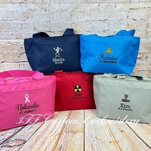 May include: Five personalized tote bags with different designs and names. The bags are made of a canvas material and have a light blue, pink, gray, red, and navy blue color.