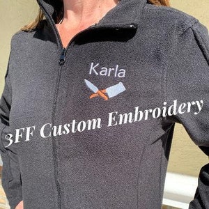 May include: Black fleece jacket with a zipper closure and a white embroidered design of crossed knives with the text "Karla 3FF Custom Embroidery".