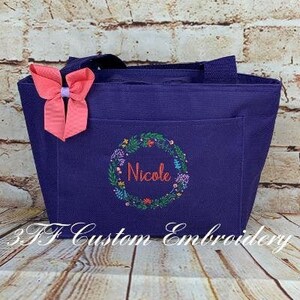 May include: A purple tote bag with a floral wreath design and the name "Nicole" embroidered in the center. A pink bow is attached to the bag.