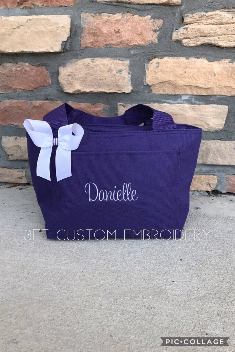 May include: A purple tote bag with a white bow and the name "Danielle" embroidered on the front. The bag is sitting on a concrete surface. 3FF CUSTOM EMBROIDERY is printed on the bottom of the bag.